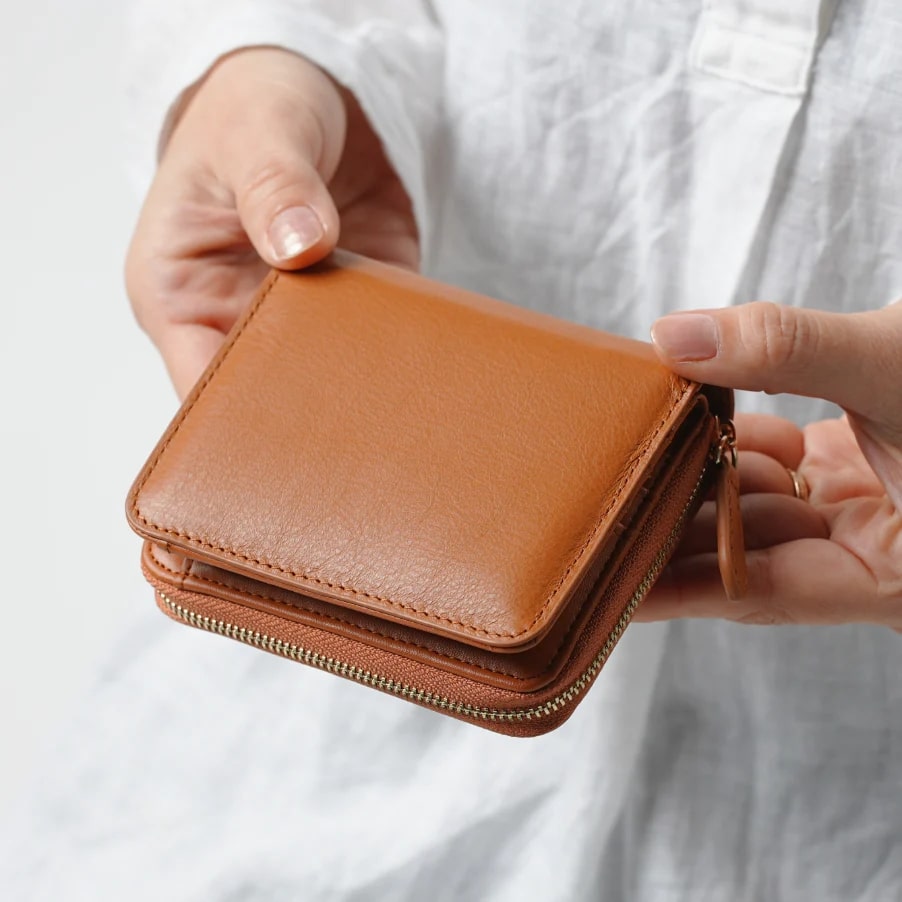 ムラ(mura）イタリア製ナッパーレザー　スキミング防止機能付きラウンドファスナー二つ折り財布（キャメル）を持つ女性