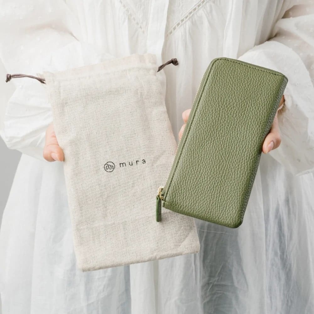 ムラのイタリアンレザー薄型L字ファスナー長財布（グリーン）と専用布袋を持つ女性